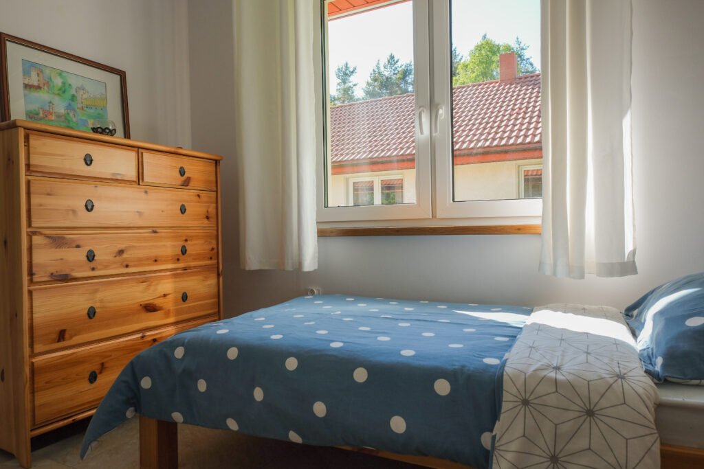 Pokój z pojedynczym łóżkiem, drewnianą komodą i oknem z widokiem na dachy.