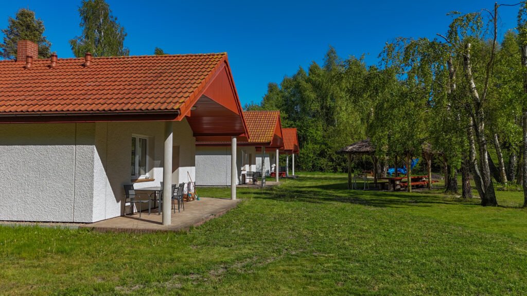 Rząd parterowych domków letniskowych z czerwonymi dachami i zadaszonymi tarasami, stojących na dużej zielonej polanie w otoczeniu drzew brzozowych; w tle widoczna altana z drewnianymi ławkami i stołami, idealna na wspólne grillowanie lub odpoczynek na świeżym powietrzu.