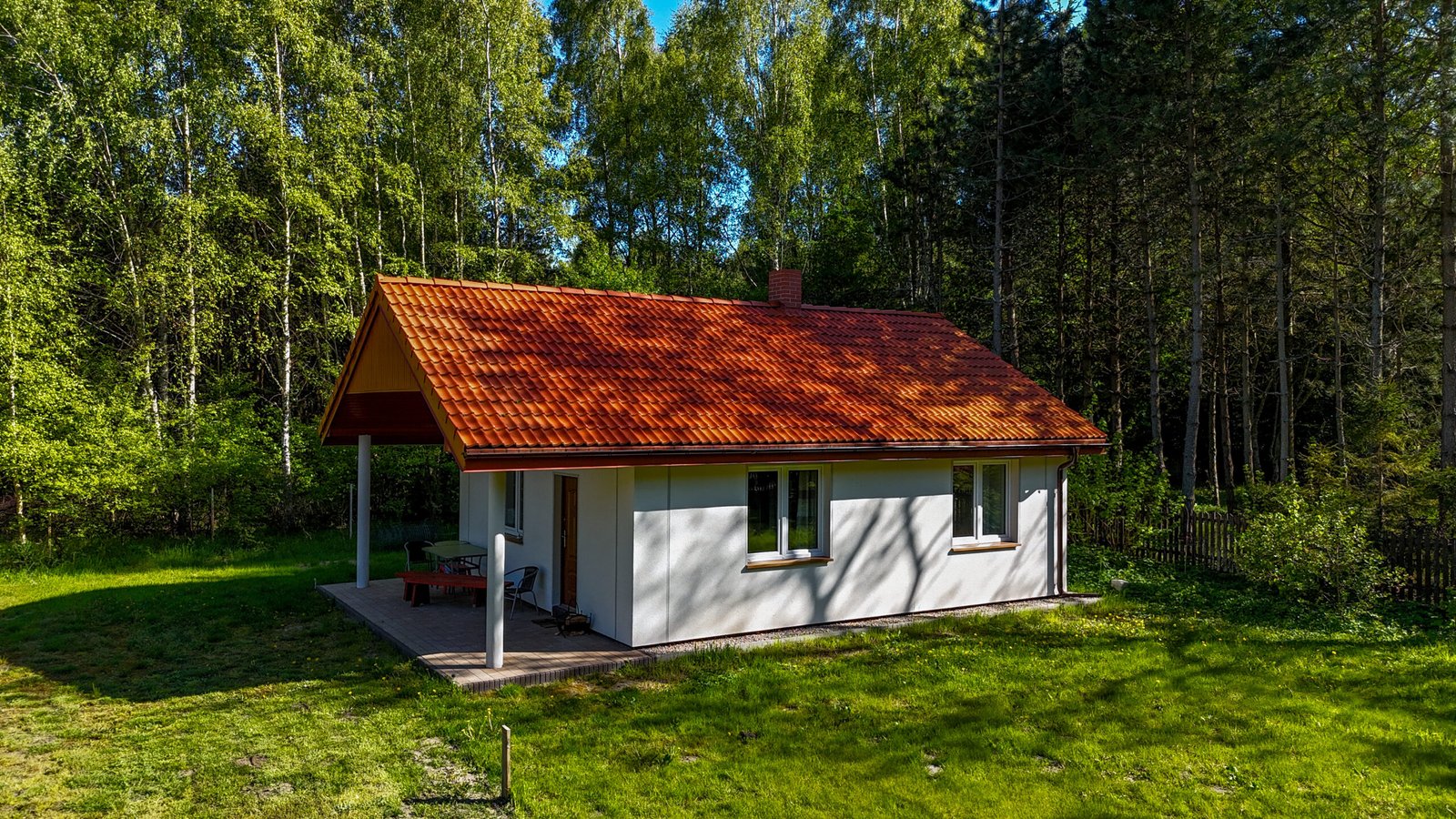 Biały domek z ceglanym dachem i werandą, idealnie wkomponowany w leśne otoczenie.