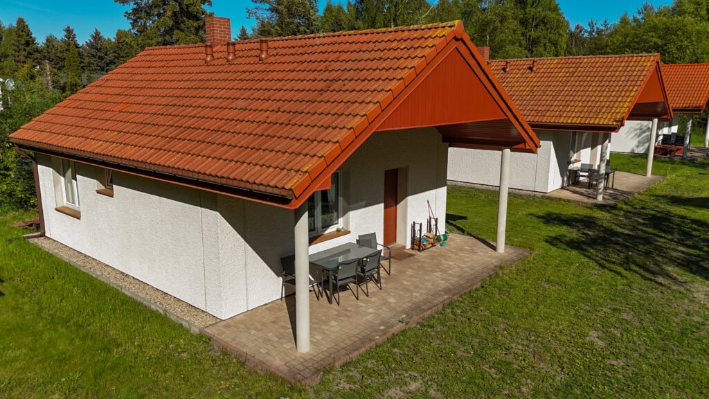 Wejście do domku z zadaszonym tarasem i zestawem ogrodowym, otoczone trawnikiem.